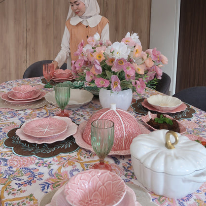 A lifestyle scene showing a woman in a white headwrap and orange top posing next to a beautifully decorated table with pink floral patterns, ceramic pumpkins, and a flower bouquet in a modern home interior. heas